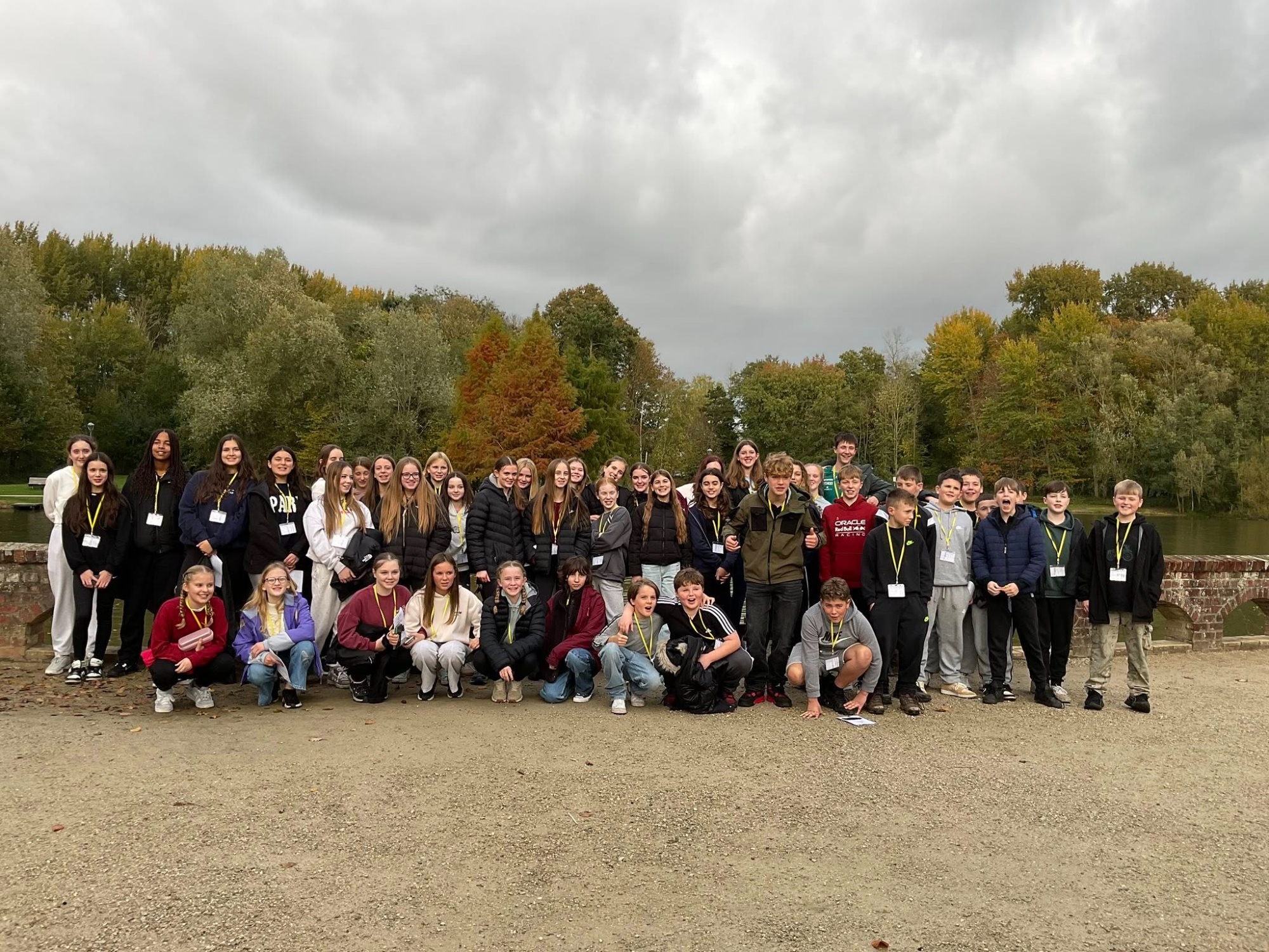A wide, landscape photo showing a large group of Year 8 pupils from the College posing together outdoors during their battlefields trip in Belgium. The group is standing on a gravel path in front of a calm lake, with a low brick bridge visible to the left. The background is filled with lush green and autumnal orange trees under a soft, overcast sky. The pupils are dressed in casual travel clothes and lanyards, with many smiling and some making friendly gestures toward the camera