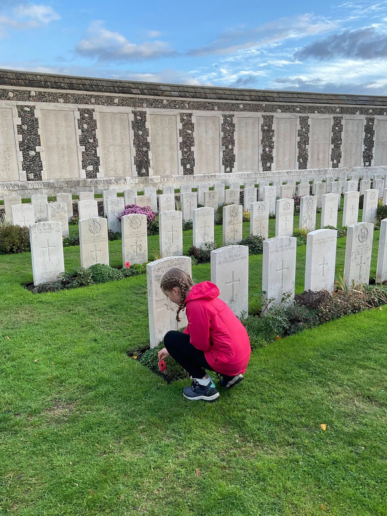 A pupil from the College wearing a bright pink jacket kneels to respectfully place a poppy at a white headstone in a Belgian war cemetery.