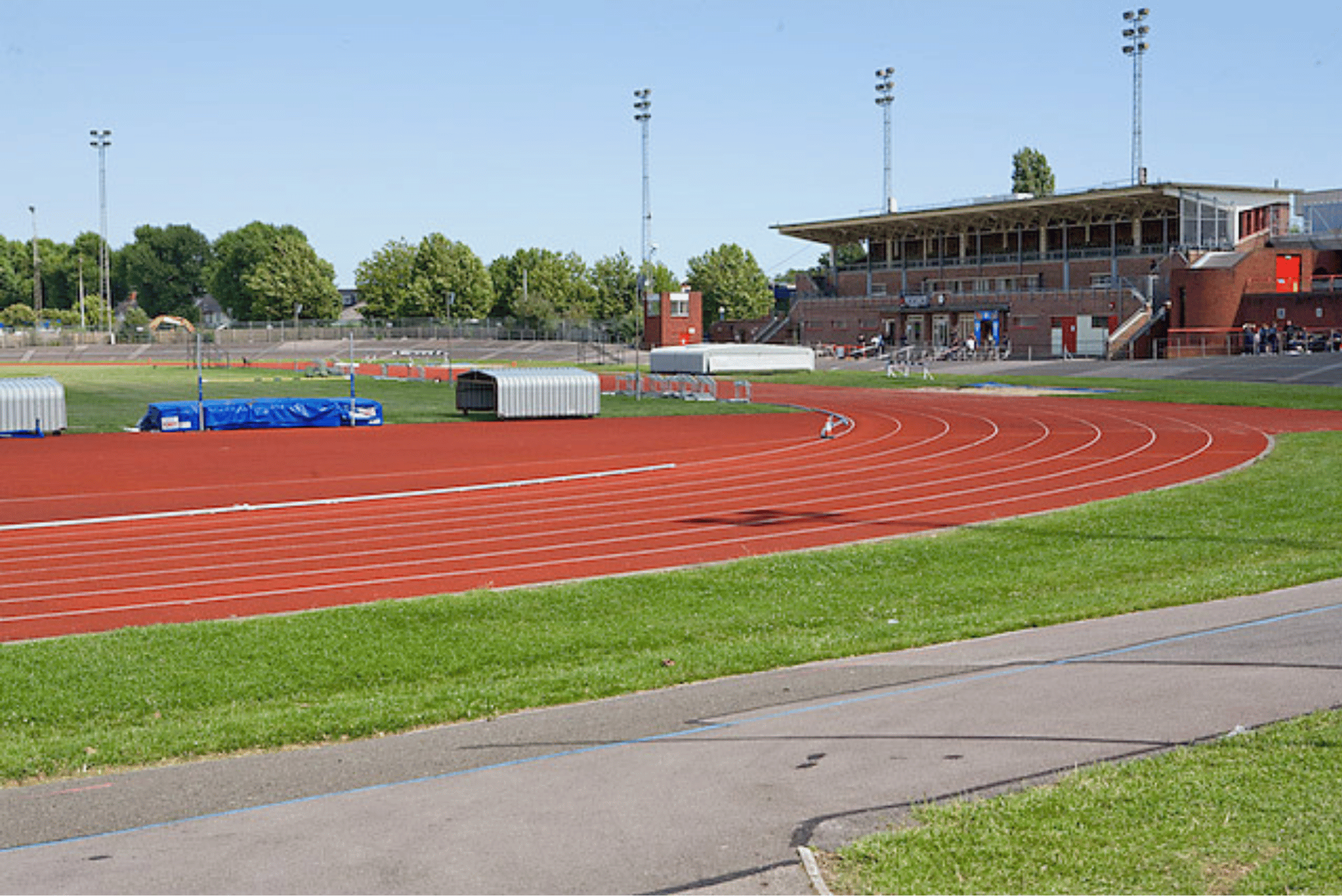 Wide view of an athletics stadium featuring a red running track curving around a grass infield. High jump equipment, storage units and athletics facilities are visible around the track. A large grandstand building stands in the background alongside floodlight towers and surrounding trees.