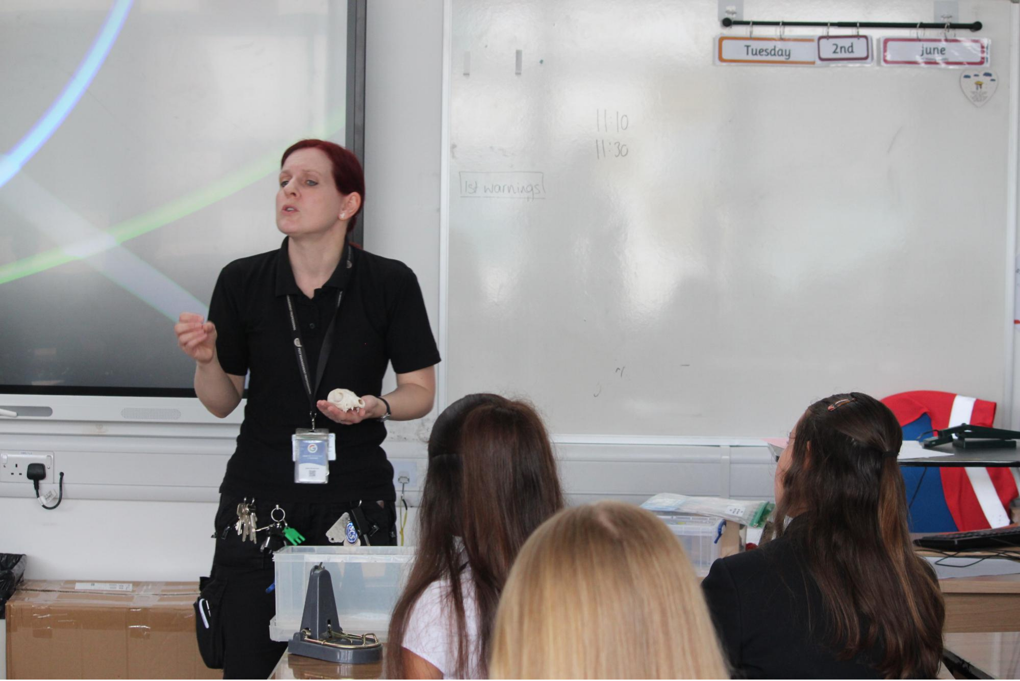 A gust speaker stands at the front of a classroom speaking to a group of pupils seated at desks. She is holding an object in one hand while presenting, with a large interactive screen and whiteboard behind her. Several pupils are visible from behind, listening to the speaker. The classroom contains storage boxes and teaching equipment. The image was taken during Swanmore College's Year 8 Meet the Employers careers event.