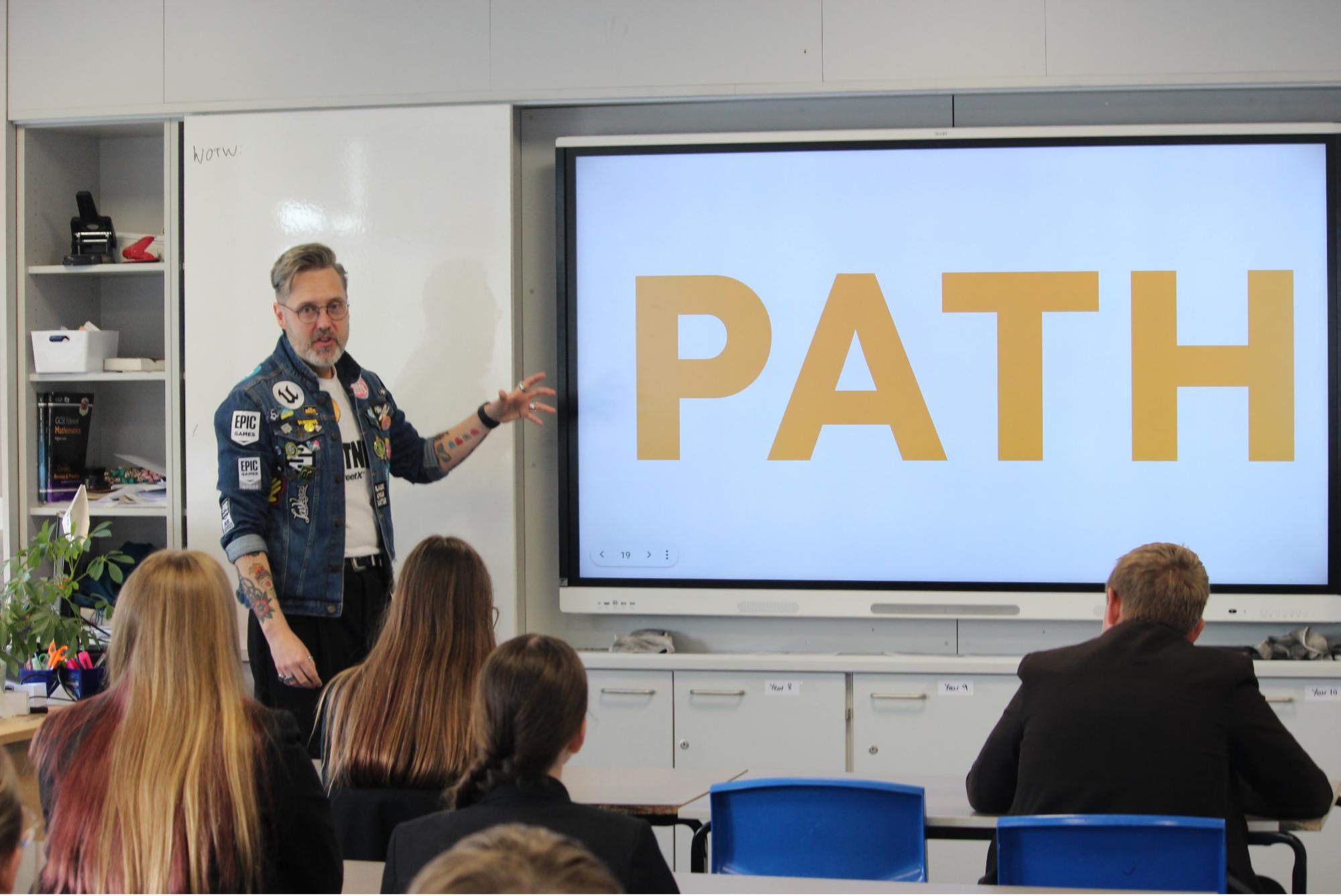 A guest speaker stands beside a large presentation screen displaying the word "PATH" in large yellow letters. The speaker is gesturing while addressing a classroom of Year 8 pupils seated at desks. The speaker is wearing a denim jacket decorated with badges and patches. Pupils are visible from behind, facing the front of the room and listening to the presentation. The image was taken during Swanmore College's Meet the Employers careers event.