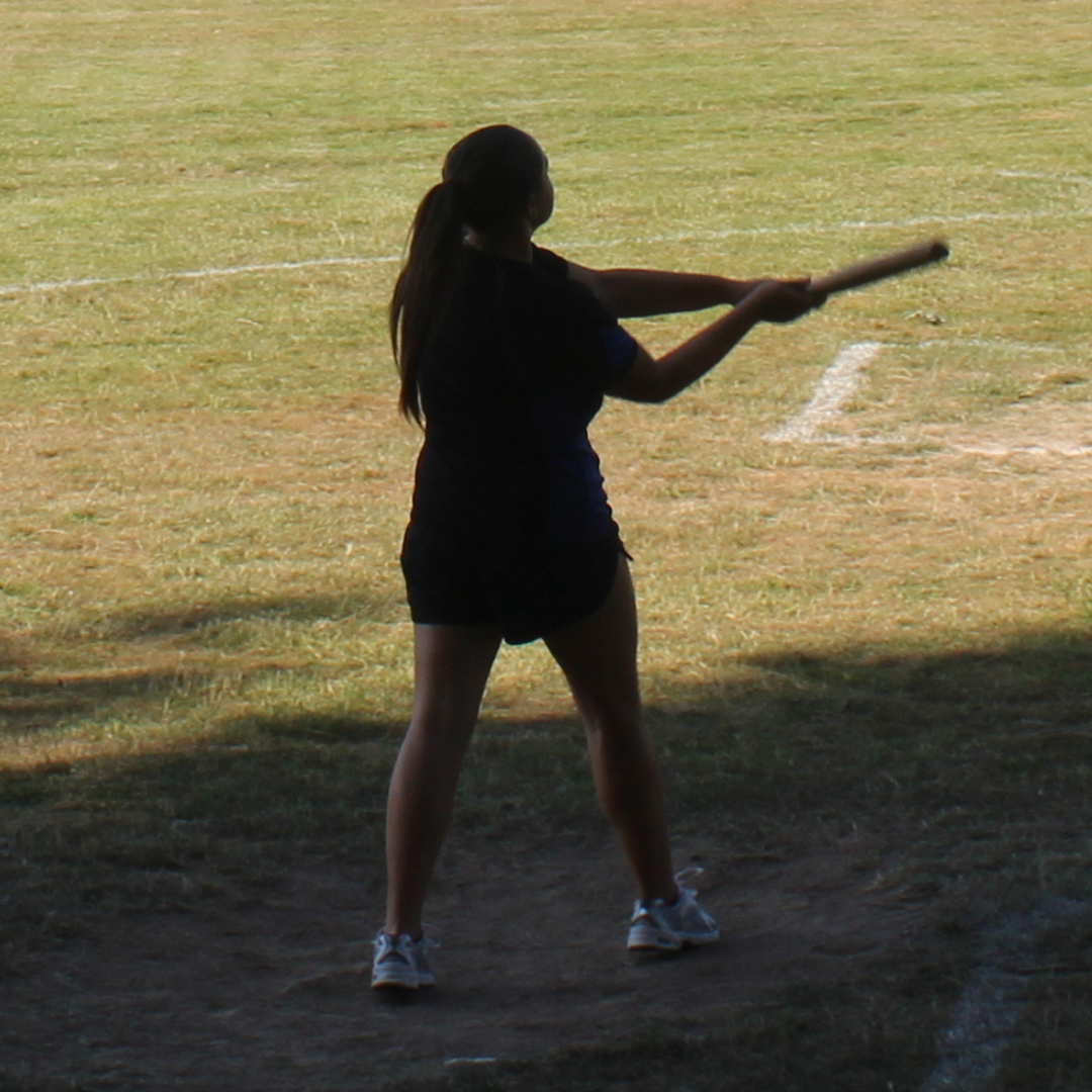 A pupil wearing Swanmore College PE kit stands on a marked grass activity area holding a rounders bat after completing a swing. The pupil is photographed from behind in partial shade, with the sports field and boundary markings visible in the background.