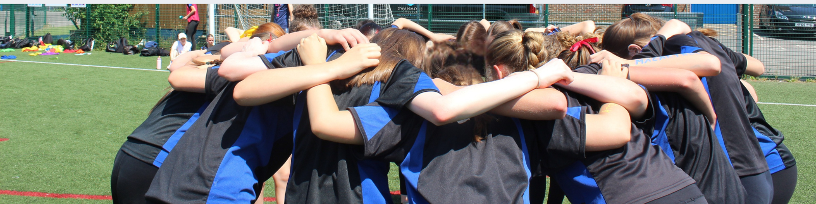 Panoramic close-up of a team huddle on the artificial pitch. Pupils wearing Swanmore College PE kit stand with their arms around one another before taking part in an activity.