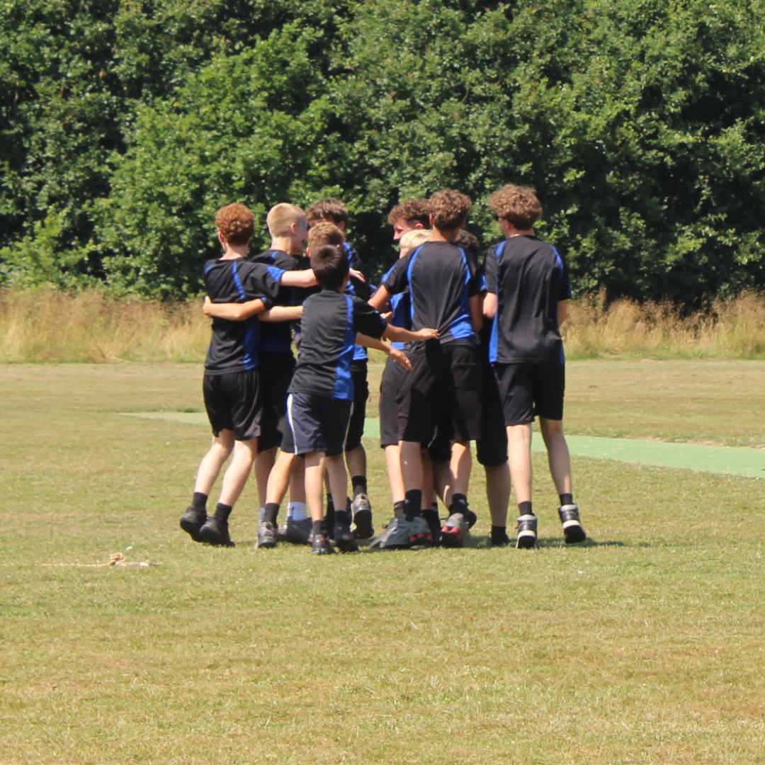 Group of pupils in black and blue Swanmore College PE kit standing in a close huddle with their arms around one another on the grass field. Trees form the background.