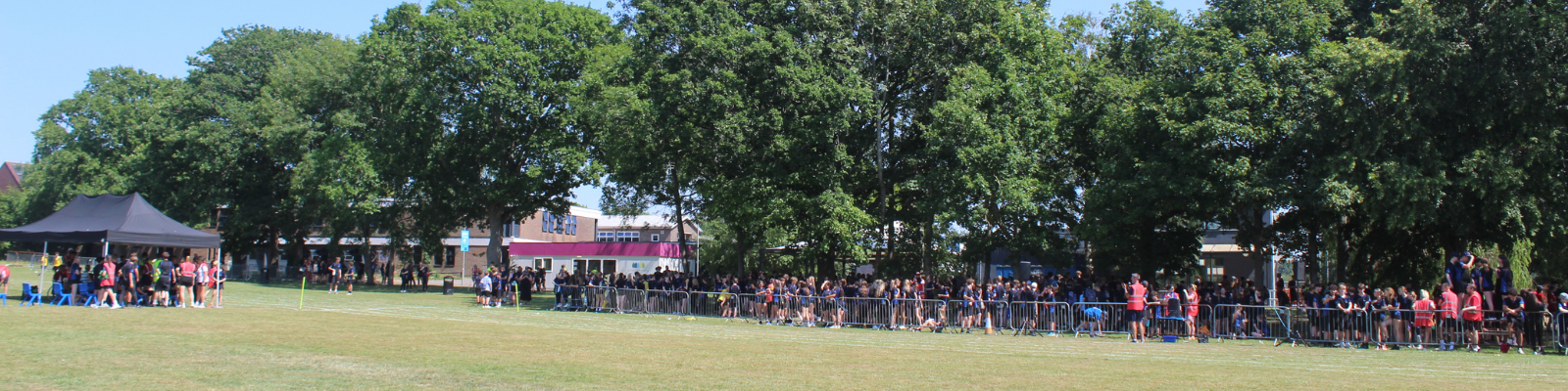 Panoramic view across the Sports Day field showing pupils gathered behind barriers and around activity areas. Event tents, staff, pupils and mature trees surround the competition area. Wide shot of two teams competing in a tug of war on the sports field. Pupils are pulling on opposite ends of the rope while staff supervise from the centre. Spectators are seated and standing behind the competition area.