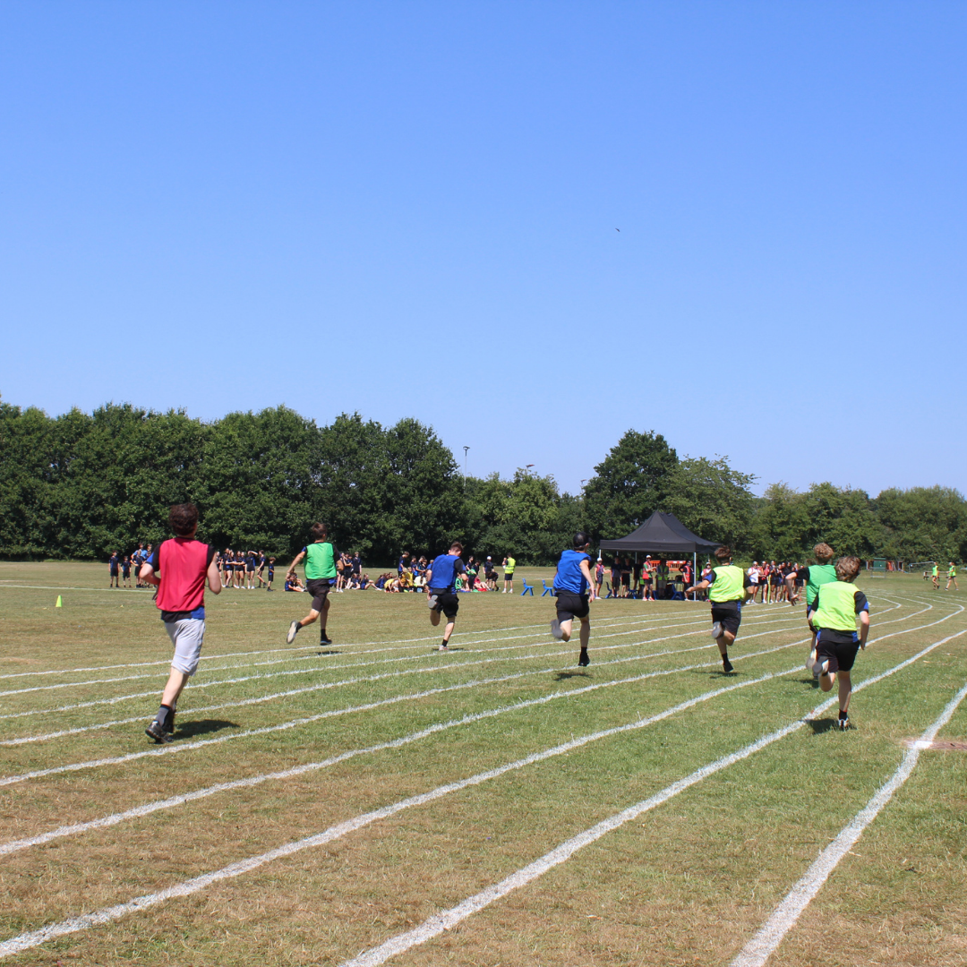Wide view of a sprint race in progress on the athletics track. Pupils wearing coloured house bibs run in marked lanes towards the finish while other pupils and staff watch from the field.
