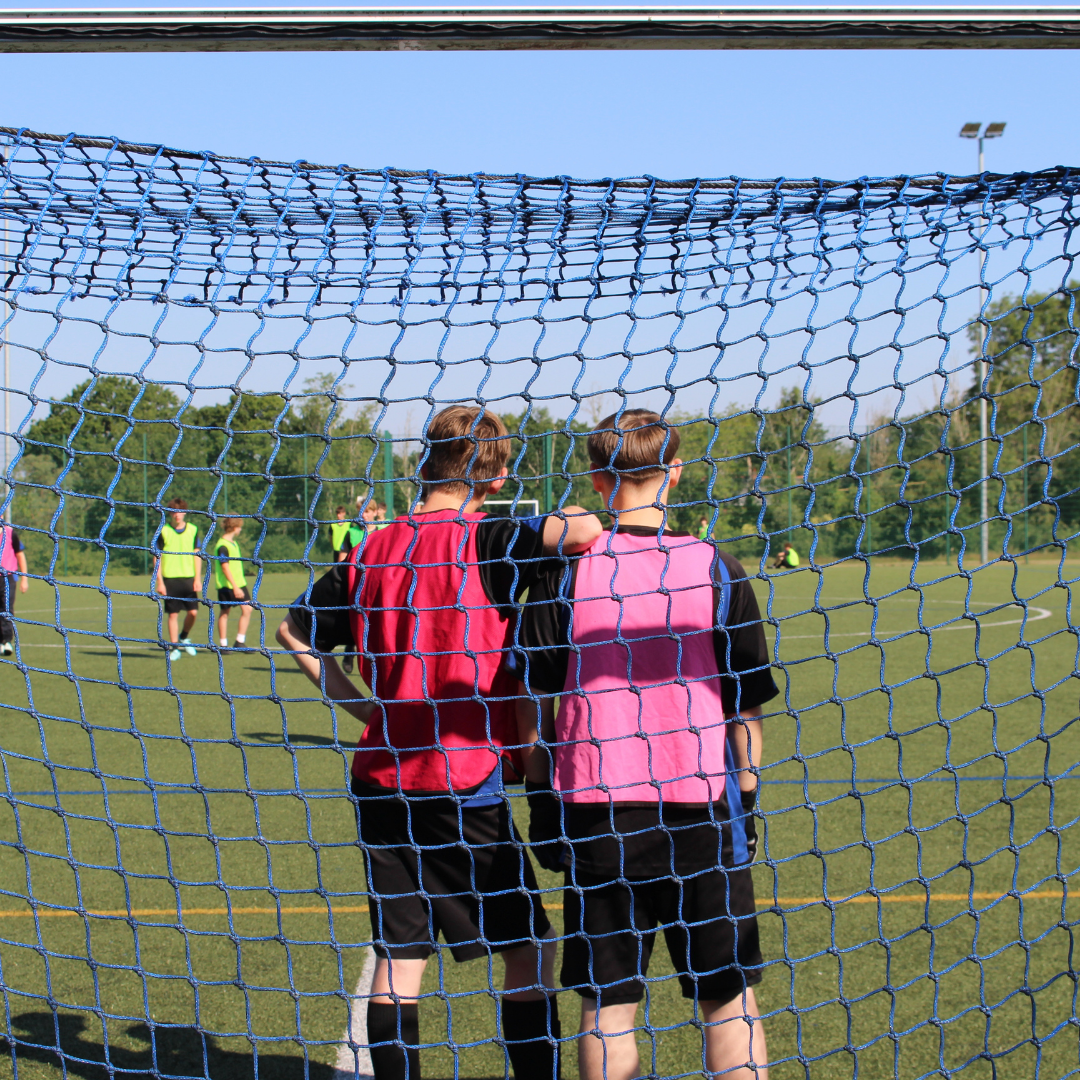 View through a football goal net showing two pupils wearing pink bibs standing with their arms around one another while watching play on the artificial pitch. Other pupils are visible in the background.