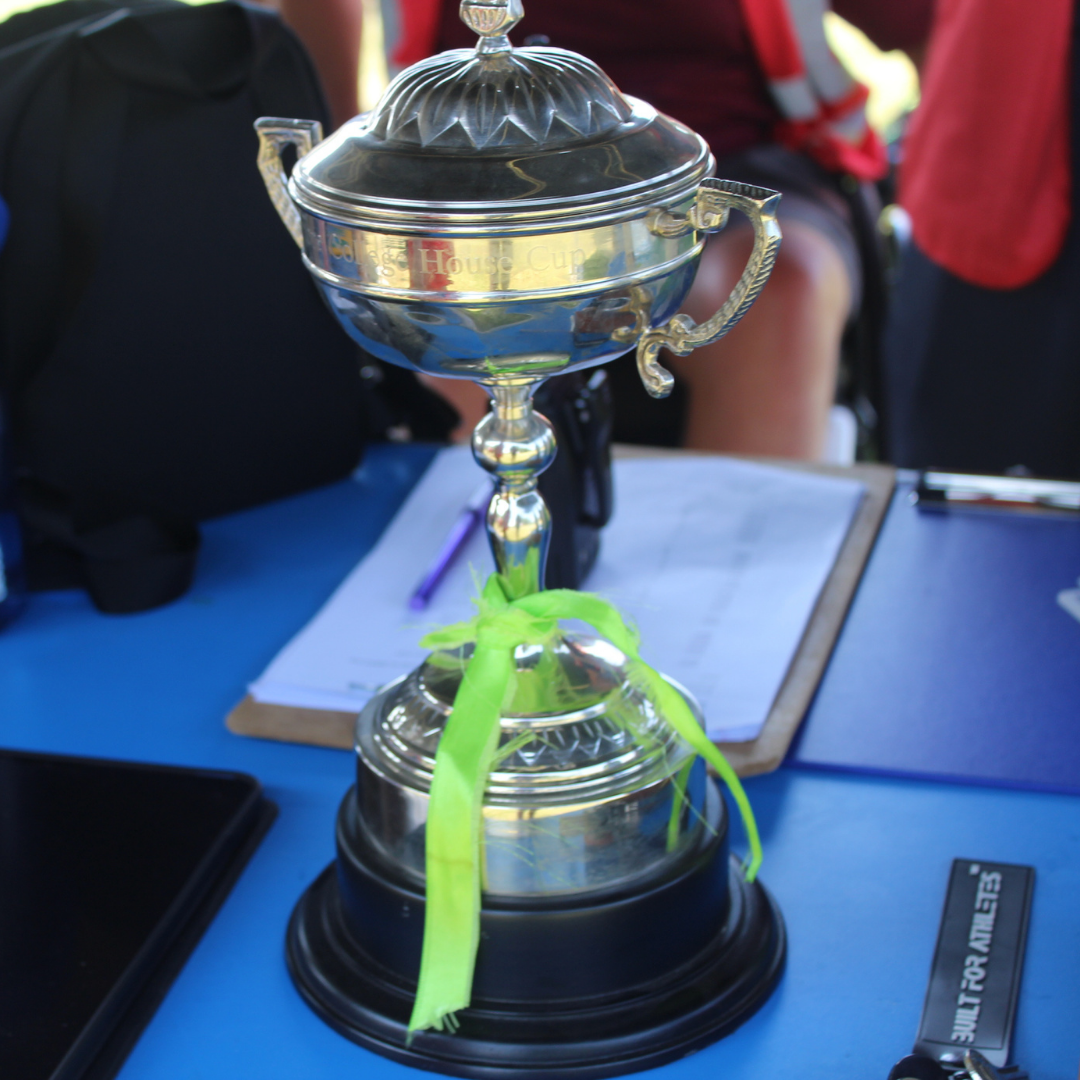 Close-up of the Sports Day House Cup trophy displayed on a blue table. A green ribbon is tied around the base of the silver trophy, with a clipboard and event equipment visible behind it.