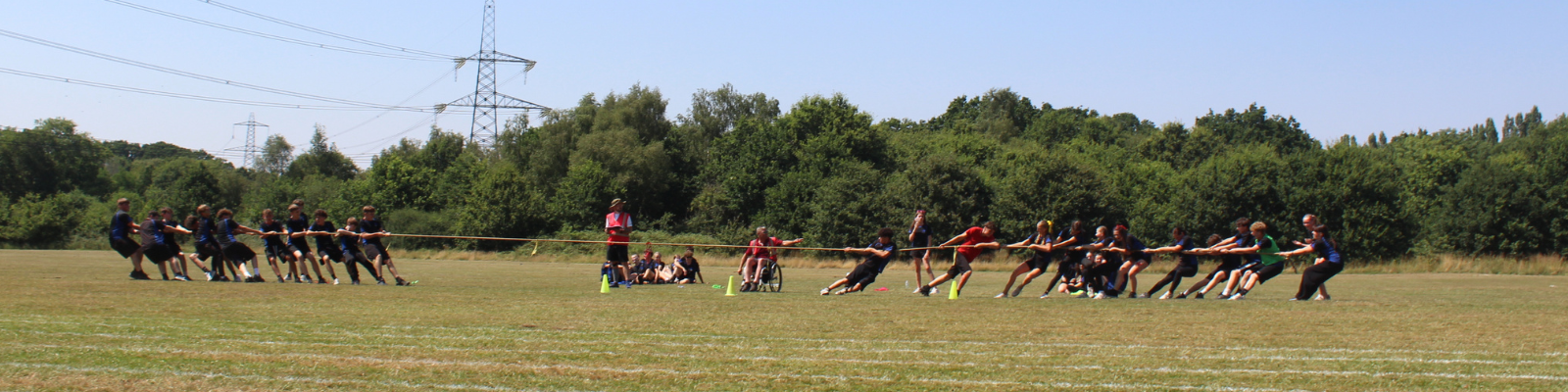Wide shot of two teams competing in a tug of war on the sports field. Pupils are pulling on opposite ends of the rope while staff supervise from the centre. Spectators are seated and standing behind the competition area.