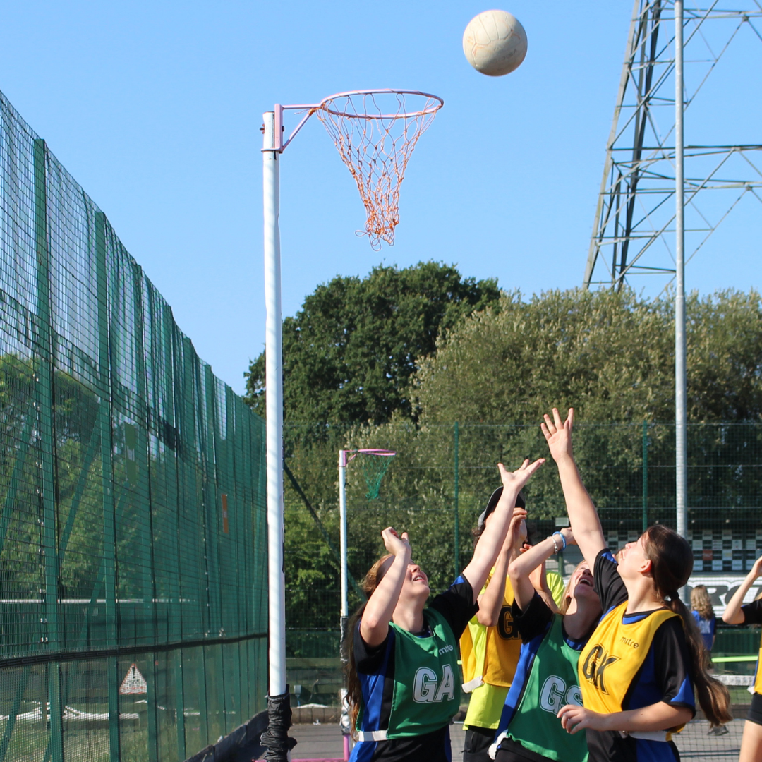 Close-up of a netball game in progress. Four pupils wearing green and yellow bibs reach towards a netball as it approaches the hoop and net on an outdoor court.