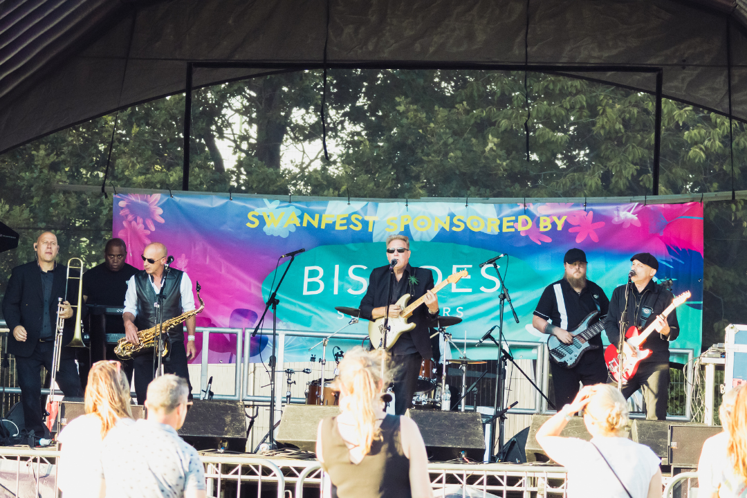Wide shot of the Rats in the Kitchen tribute band performing on the Swanfest main stage. Six band members are visible with brass instruments, guitars, bass guitar, keyboard and drums. Audience members are standing in front of the stage.