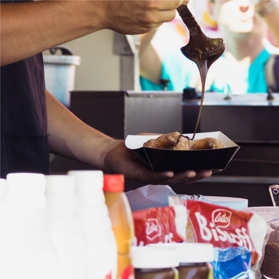 Close-up of a food vendor pouring chocolate sauce over a tray of cinnamon-coated doughnuts. Ingredients and serving containers are visible on the counter in the foreground.