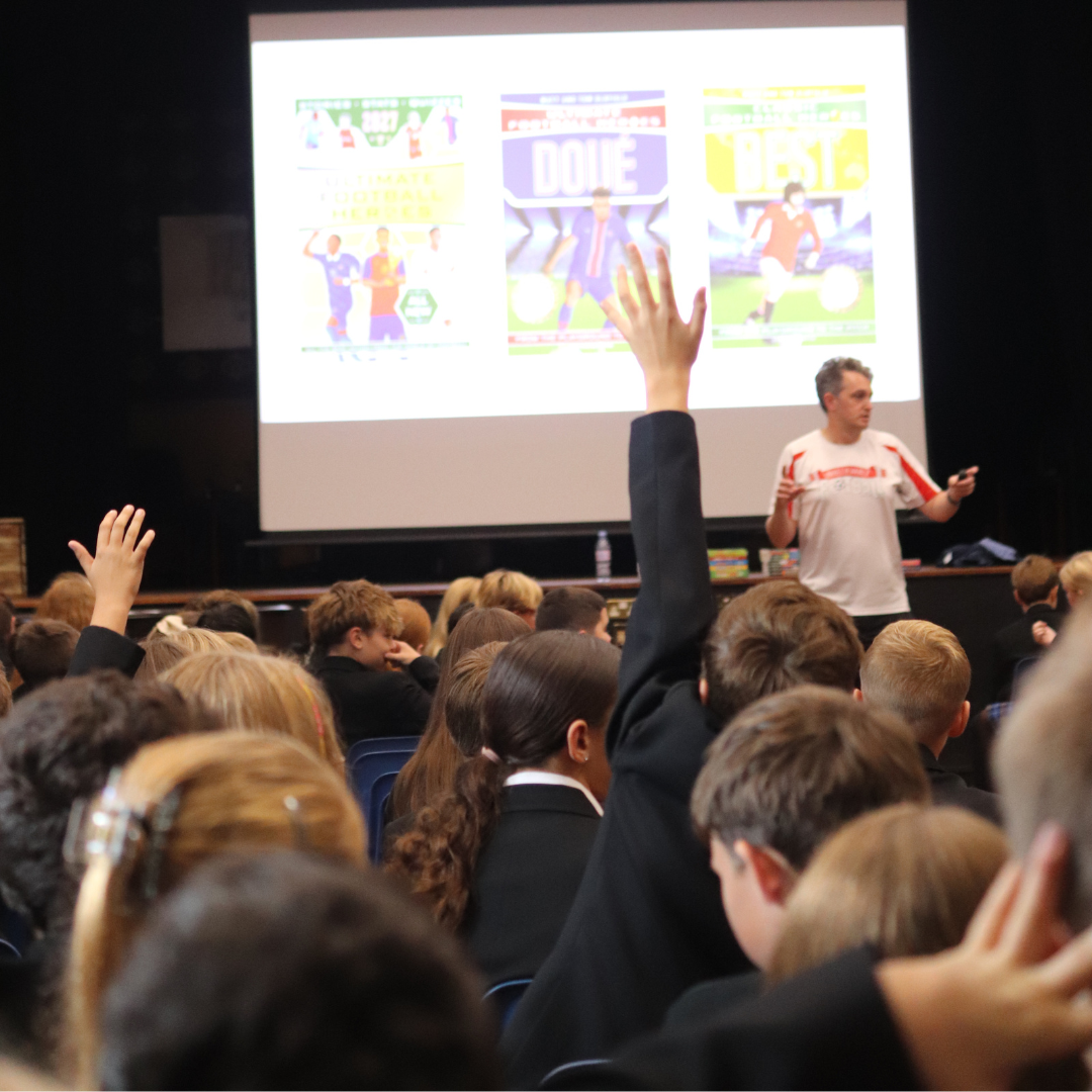 Pupils raising their hands to ask questions during author Matt Oldfield's talk in the hall, with book covers projected on the screen behind him.