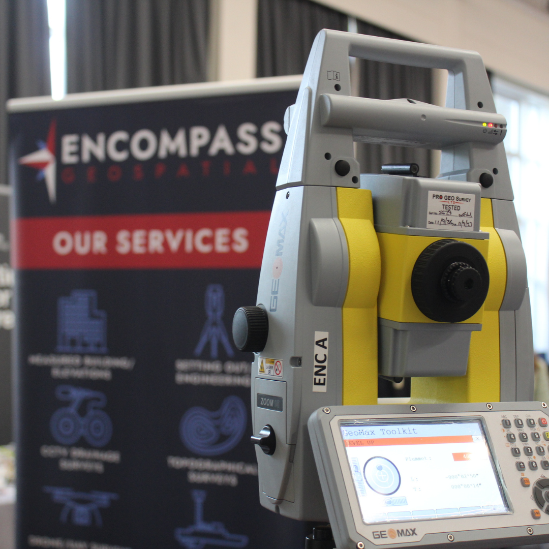 A GeoMax total station survey instrument set up on display in front of an Encompass Geospatial exhibition banner.