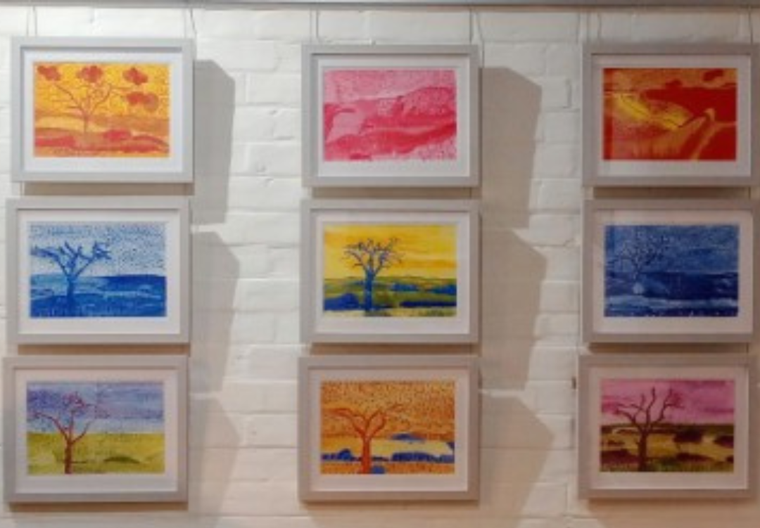 Image shows nine pieces of colourful, framed artwork on display against a white brick wall.