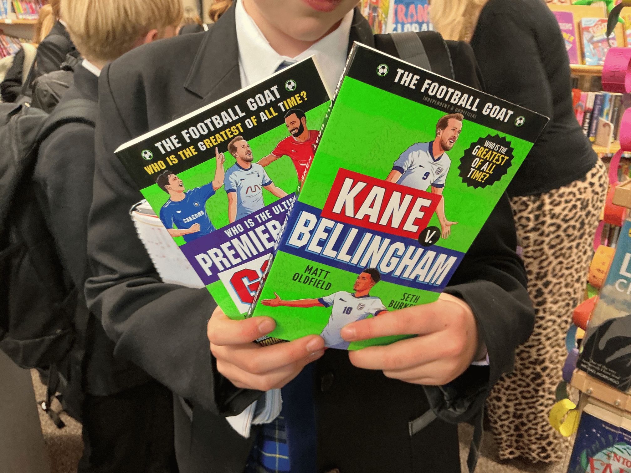 A pupil holding copies of books by Matt Oldfield, including "Kane v. Bellingham" and "Who is the Ultimate Premier League GOAT?" during an author visit event.