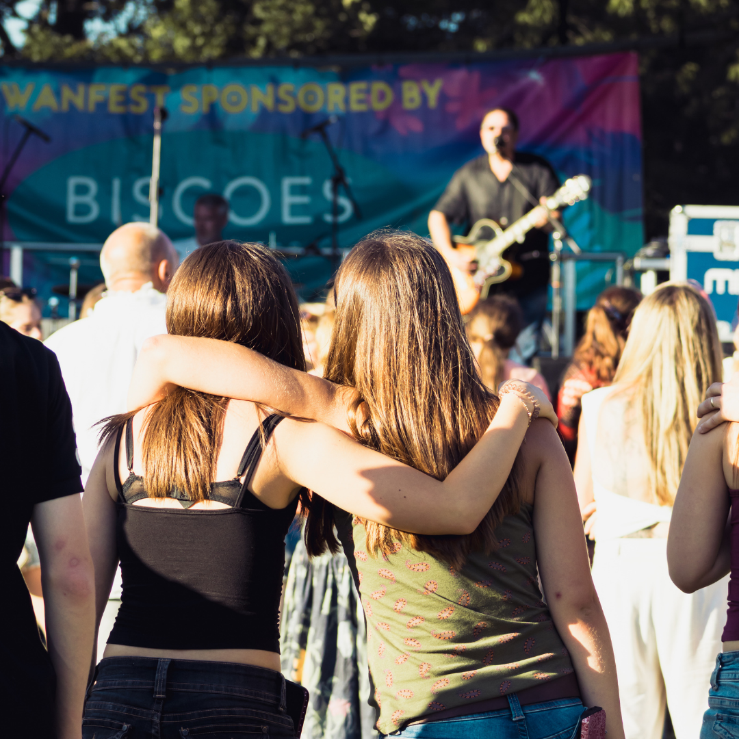 Rear view of two audience members standing with their arms around each other while watching a live performance on the main stage. A performer is visible out of focus in the background.