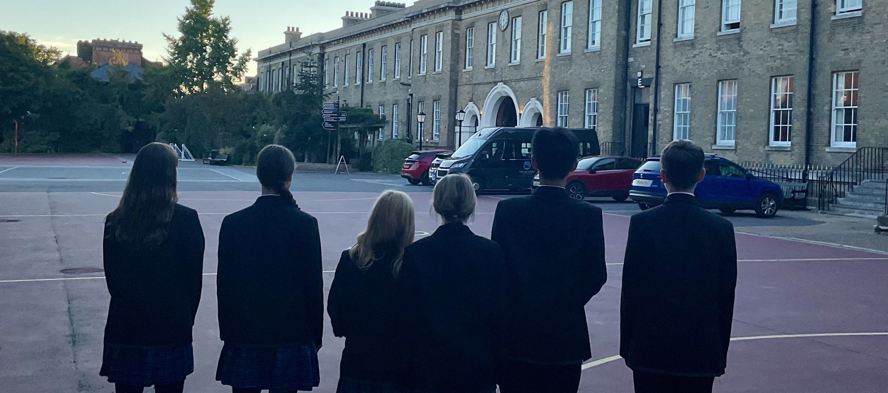 A photograph showing six Swanmore College debate team students viewed from behind, standing on a outdoor court area outside a historic school building.