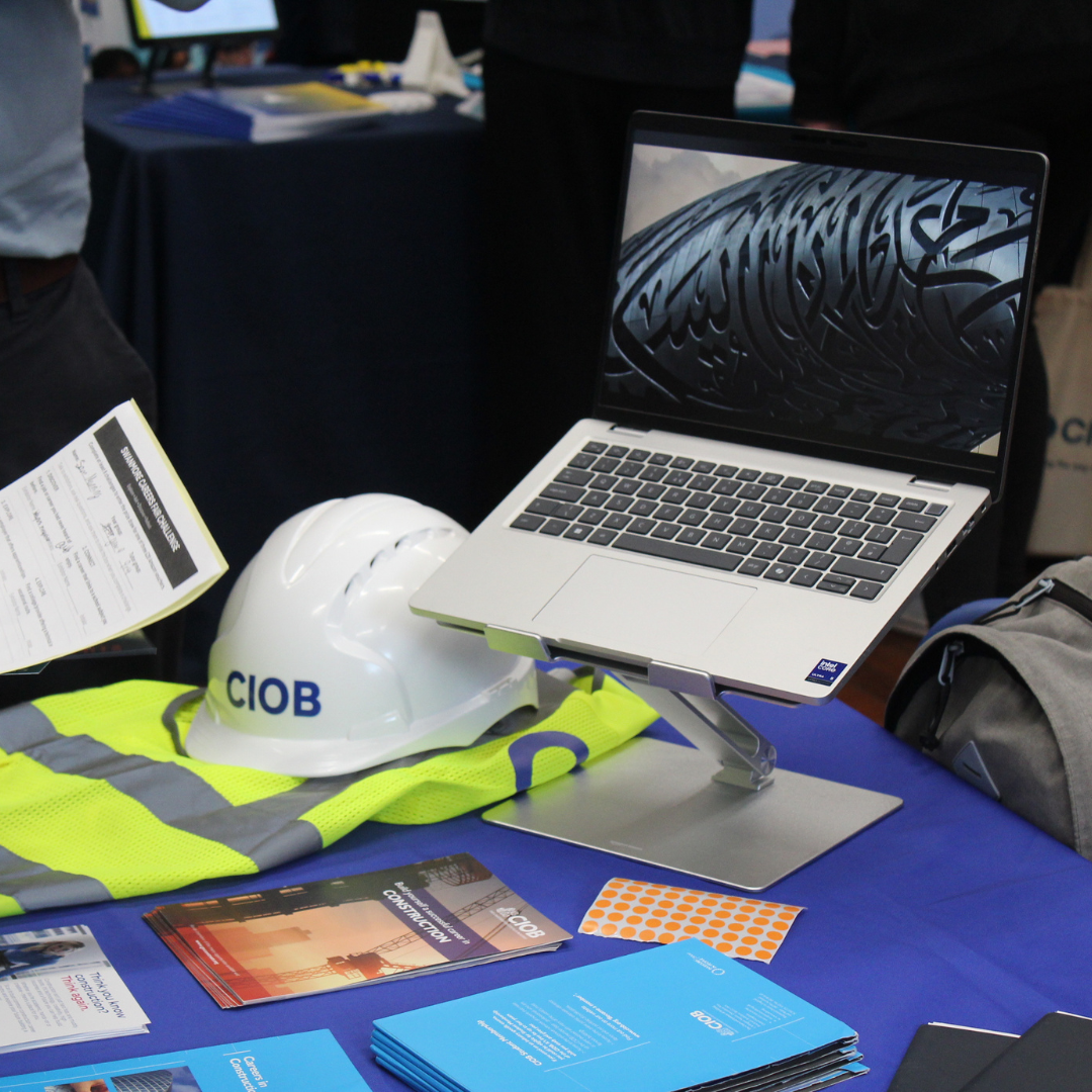 A CIOB construction display table featuring a hard hat, high-visibility vest, laptop, and promotional literature.