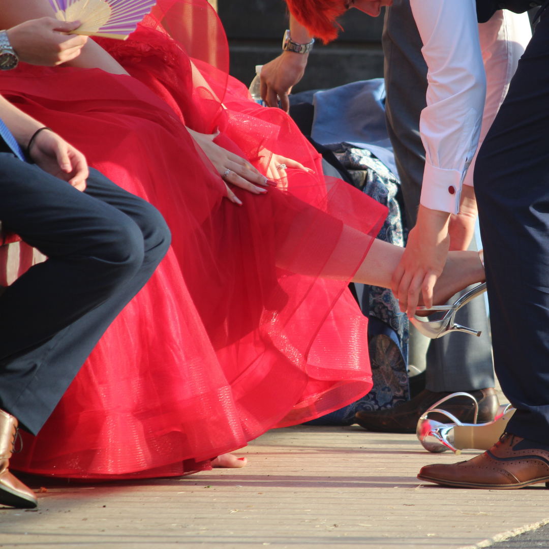 Pupil helping another pupil fasten a silver high-heeled shoe while seated.