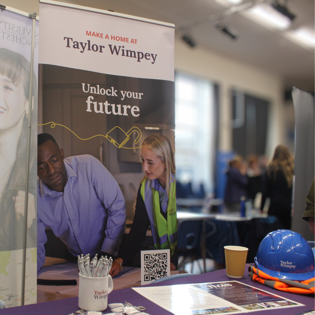 A Taylor Wimpey exhibition stand featuring a banner, hard hat, and information leaflets.