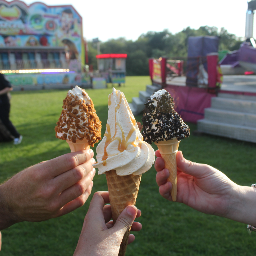 Three hands holding ice cream cones with fairground rides in the background.