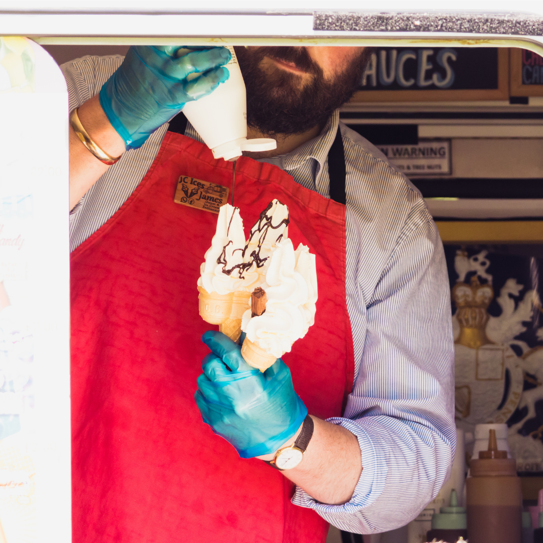 Close-up of a member of an ice cream vendor preparing two soft-serve ice creams with chocolate sauce. The vendor is wearing blue gloves, a red apron and a striped shirt inside the serving hatch of an ice cream van.