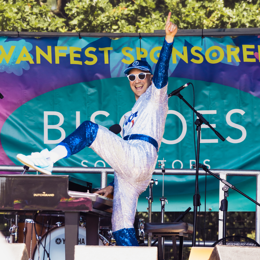 A performer dressed in a blue and silver Elton John-inspired sequinned costume poses on the Swanfest stage with one leg raised and one arm pointing upwards. A keyboard and stage equipment are visible around them.