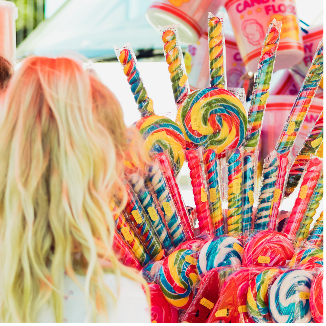 Close-up of a display of wrapped rainbow-coloured spiral lollipops and candy sticks at a confectionery stall. The back of a person with long blonde hair is visible in the foreground.