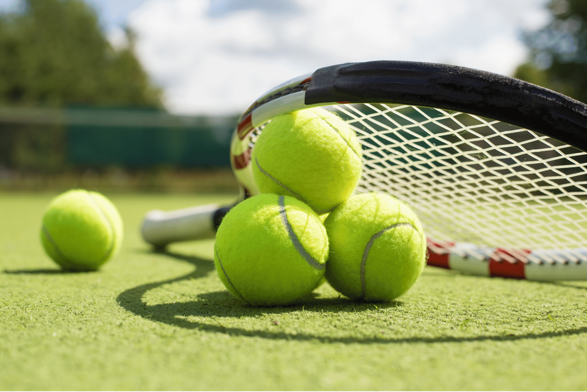 Close-up photograph of four bright yellow tennis balls resting on an outdoor tennis court beside a tennis racket. Three balls are grouped beneath the racket head, with a fourth ball positioned separately in the foreground. The racket lies on the court surface with its strings and frame clearly visible. Trees, fencing and a blue sky with clouds appear blurred in the background.
