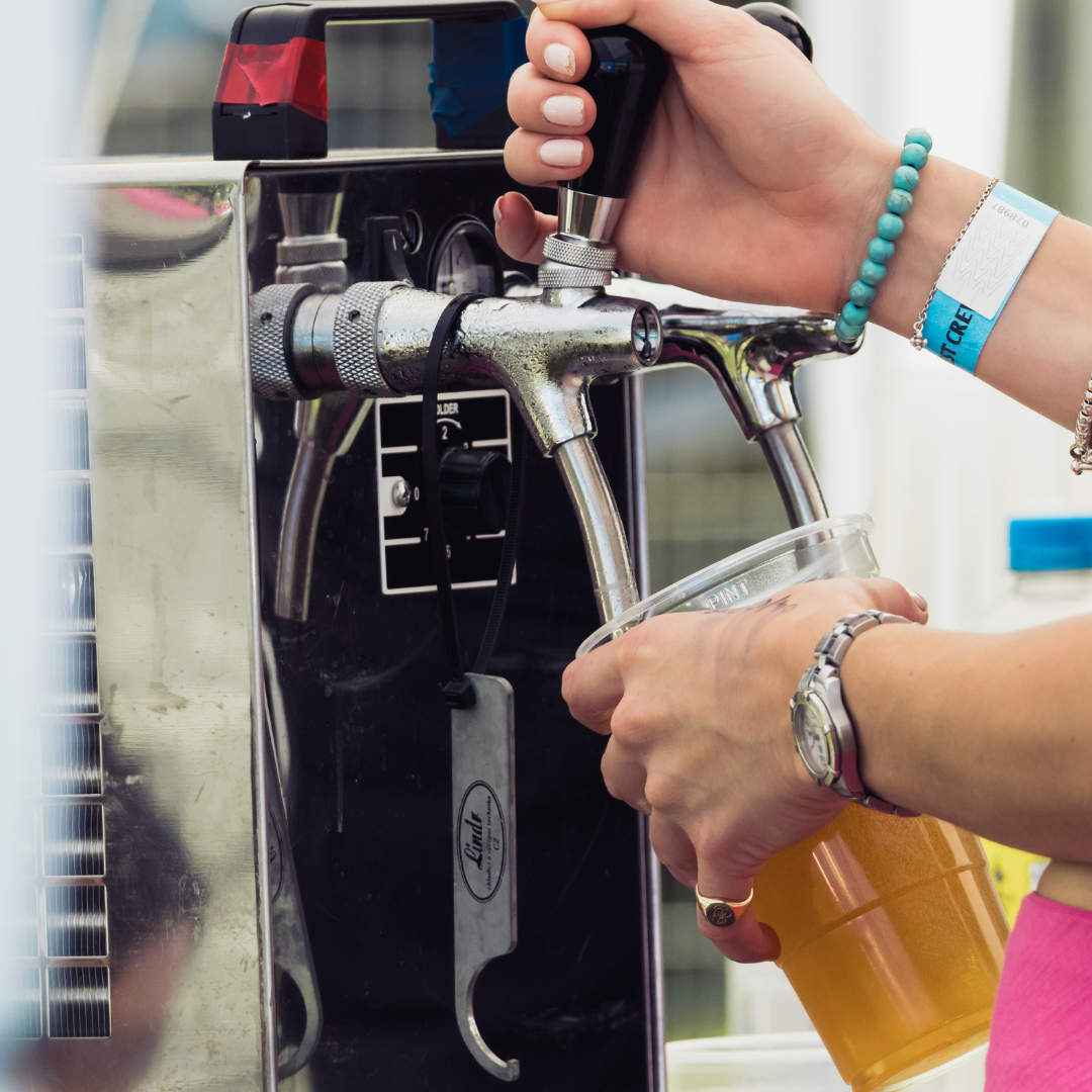 Close-up of a person pouring a drink from a beer tap into a clear plastic cup. The person's hands, wristband, bracelet and watch are visible, along with the stainless steel drinks dispenser.