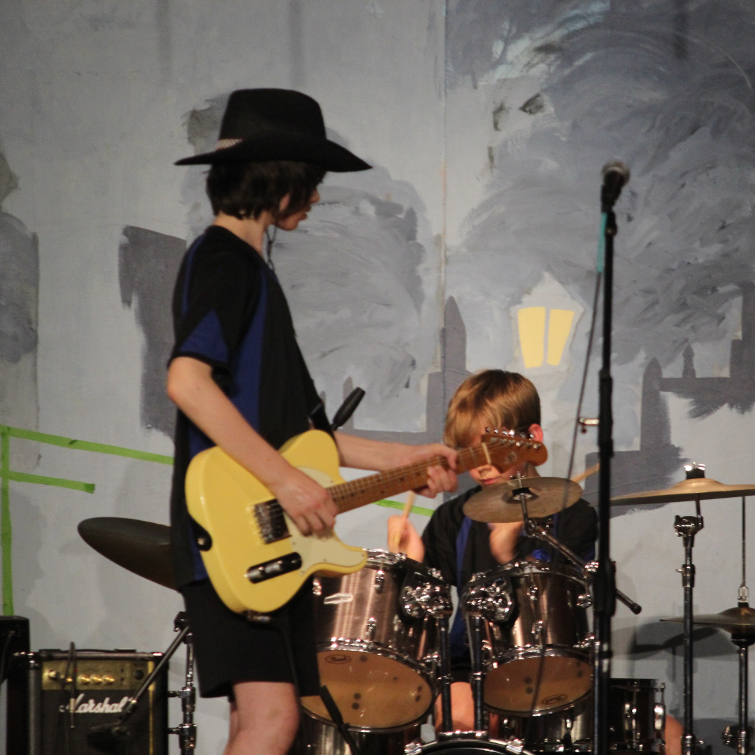 Image shows two pupils dressed in PE kit, one playing the electric guitar and wearing a hat, the other playing a drum kit