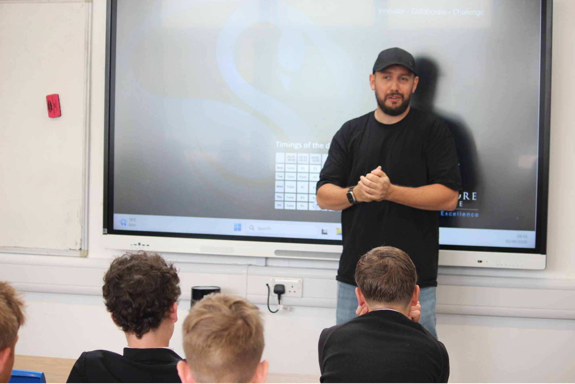 A guest speaker stands in front of a large interactive screen addressing a classroom of Year 8 pupils. The speaker is wearing a black top and cap and is gesturing while speaking. Pupils are seated facing the front of the room and listening attentively. The screen behind the speaker displays the Swanmore College desktop background. The image was taken during the Year 8 Meet the Employers careers event.