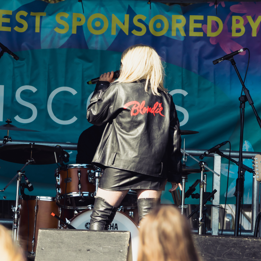 Rear view of a performer wearing a black leather jacket with "Blondie" written across the back in red lettering. The performer is holding a microphone on the Swanfest stage with drums and microphone stands visible behind.