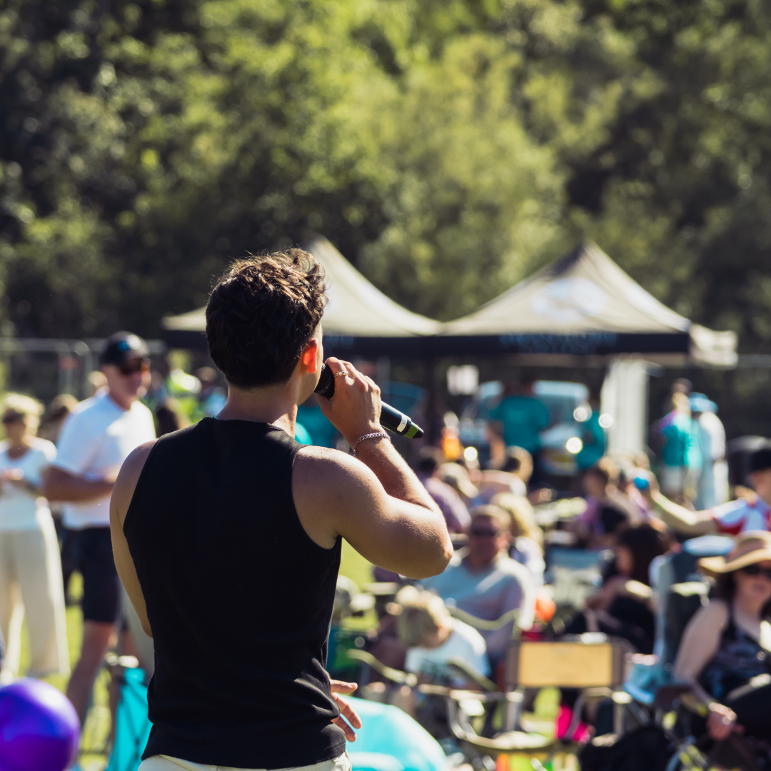 Rear view of a singer holding a microphone while performing amongst the audience. Festivalgoers are seated and standing on the grass, with event tents visible in the background.