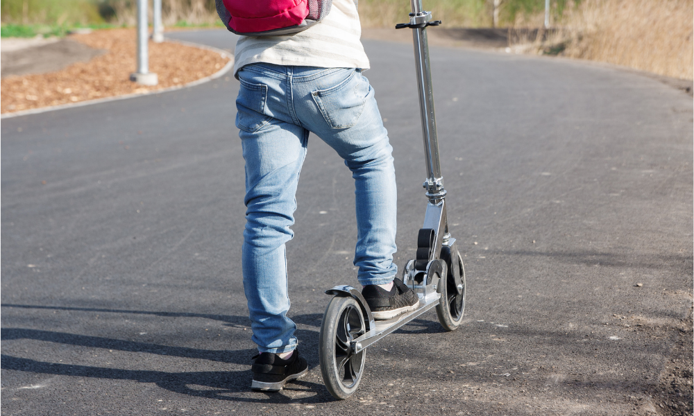 Lower body view of a person wearing jeans and trainers riding a silver kick scooter along a paved pathway.