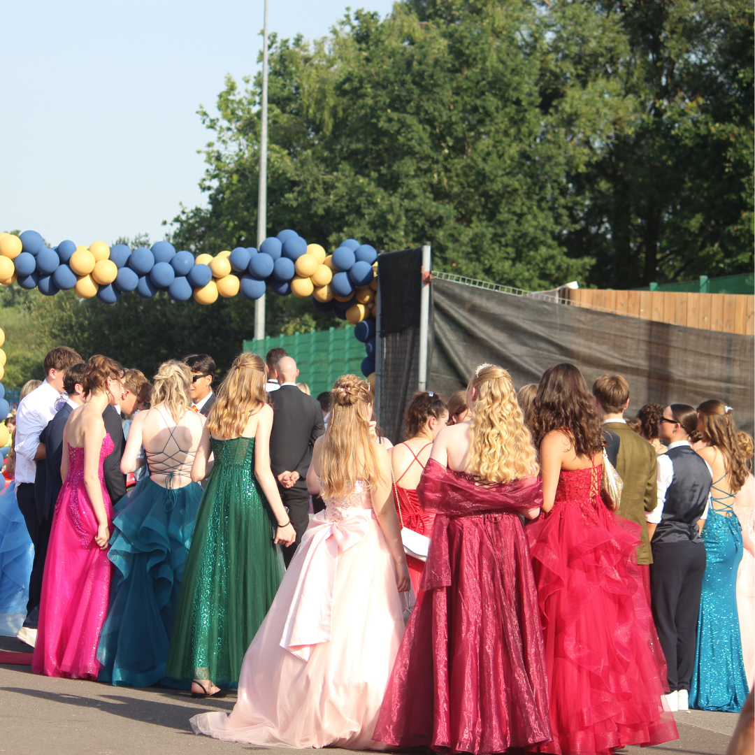 A group of pupils stands outdoors beneath a blue and gold balloon arch at a Year 11 Prom arrival area. The image is taken from behind, showing pupils gathered in formal attire, including a variety of floor-length dresses in bright pink, teal, green, pale pink, burgundy and red, alongside pupils wearing suits. Trees, fencing, a speaker and a lamppost are visible in the background. Bright afternoon sunlight casts shadows across the road surface.