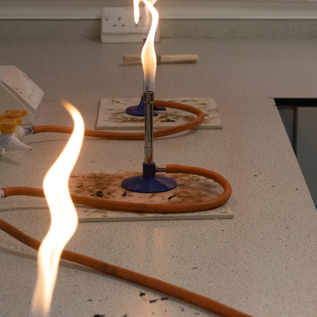 Image shows three lit Bunsen burners arranged in a row on a light grey science laboratory worktop. Each burner sits on a white heatproof mat marked with brown scorch stains, and orange gas hoses loop around the bases and connect to gas taps. Tall yellow-orange flames rise from the metal barrels, with one flame blurred in the foreground. A wall socket and laboratory equipment are visible in the background, indicating a practical science lesson.