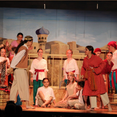 Image shows a stage performance with pupils in colourful costumes, standing and seated in character as part of a dramatic production set against a painted backdrop.