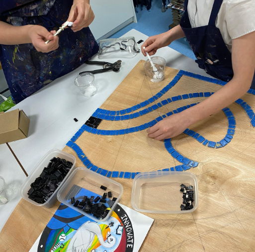 Pupils working collaboratively on a mosaic artwork, placing blue tiles onto a wooden board using adhesive and tools in an art room.