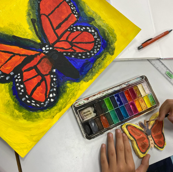 A brightly coloured painted butterfly on yellow paper, alongside a paint palette and a pupil’s hand painting a butterfly model.