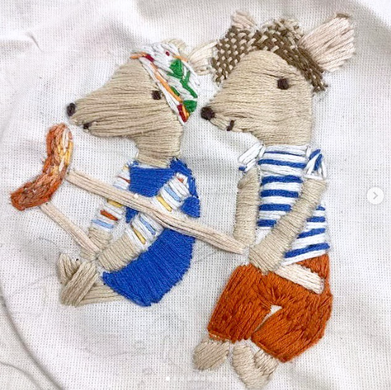 An embroidered textile artwork showing two stylised animals in clothing, stitched in thread on fabric with detailed textures and patterns.