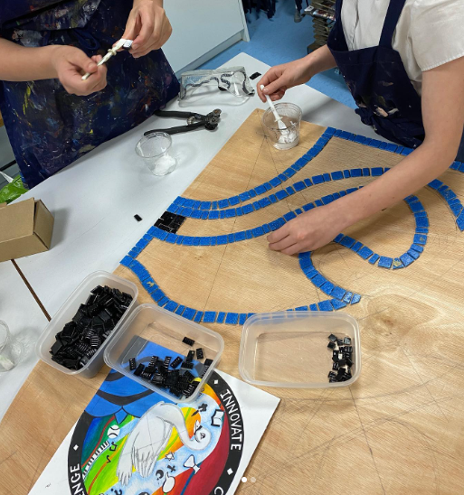 Pupils working collaboratively on a mosaic artwork, placing blue tiles onto a wooden board using adhesive and tools in an art room.