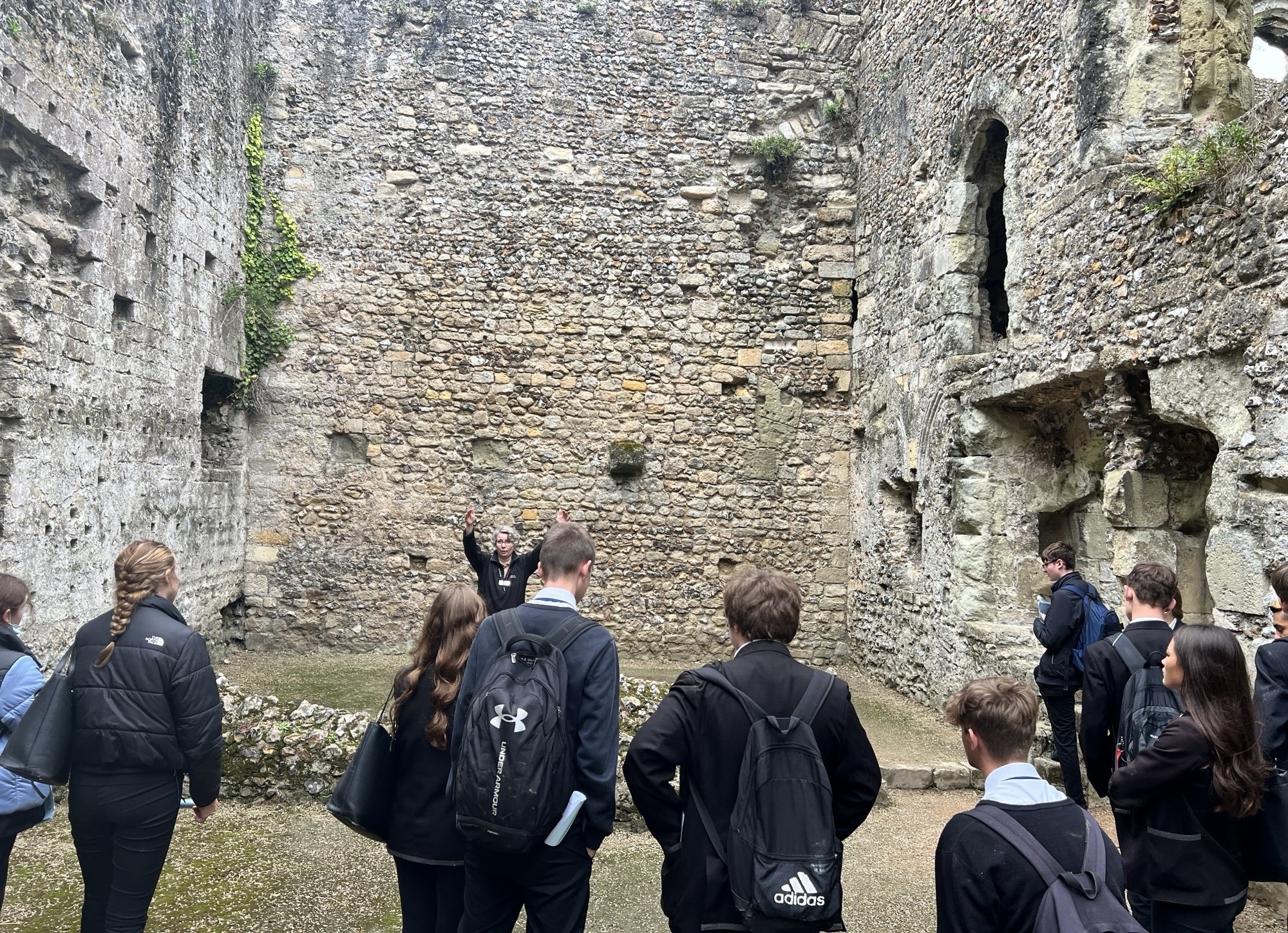 A group of pupils in school uniform and outdoor coats stand inside the high, weathered stone walls of a castle ruin. They are listening to a teacher who is gesturing with raised arms while explaining the history of the site.