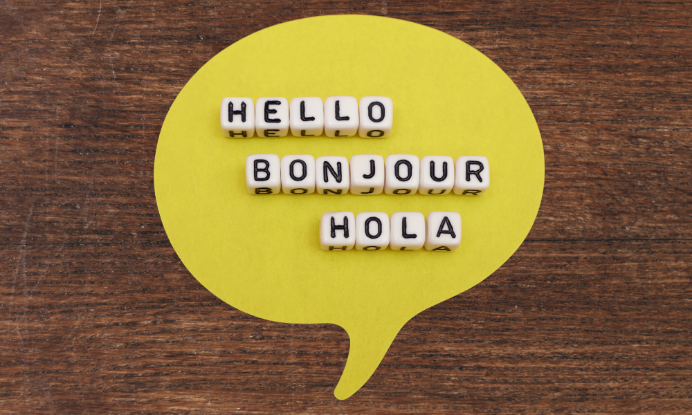 A top-down view on a rustic wooden surface featuring a bright yellow circular speech bubble. Inside the speech bubble, white letter blocks spell out the word "HELLO" in English, "BONJOUR" in French, and "HOLA" in Spanish.