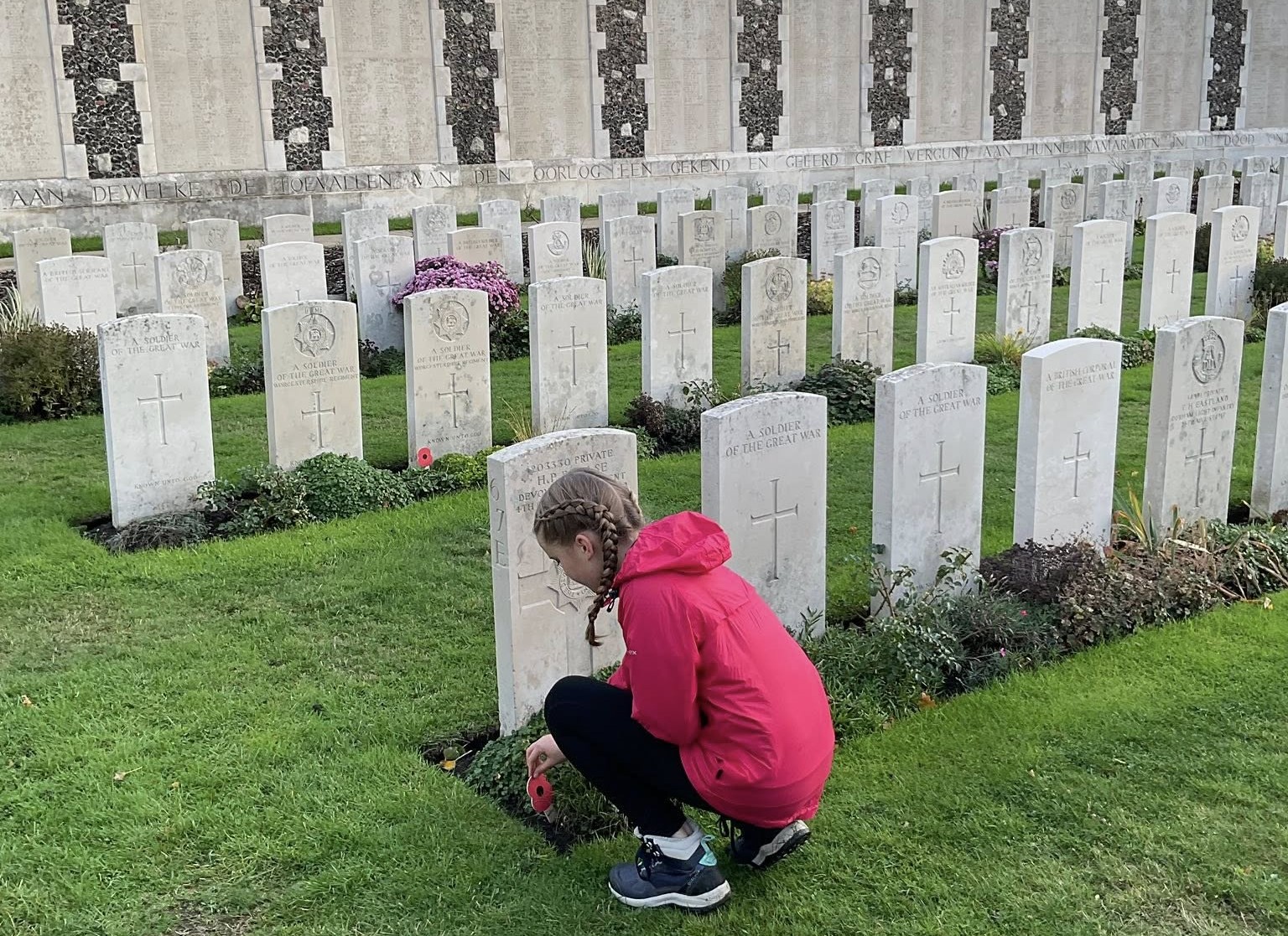 A pupil in a pink jacket kneels to place a red remembrance poppy at the base of a white headstone in a meticulously kept military cemetery. Rows of identical white graves stretch into the background toward a stone memorial wall.