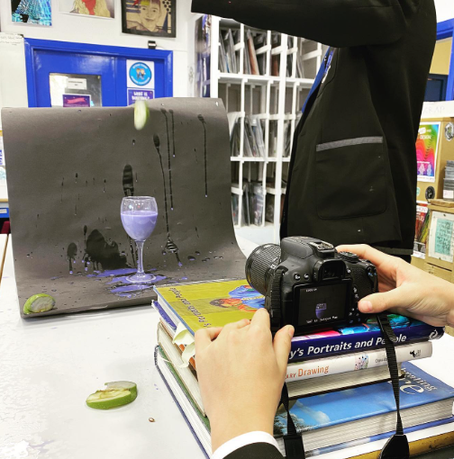 A pupil photographs a still-life setup using a digital camera balanced on art books, showing a glass, splashes of liquid, and fruit arranged against a dark backdrop in an art classroom.