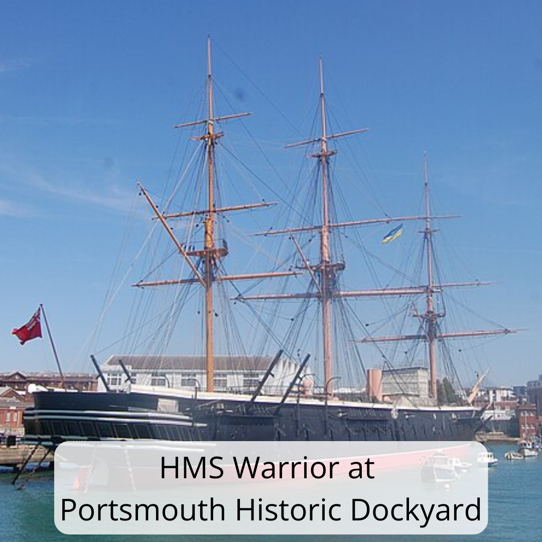 Image shows HMS Warrior moored at Portsmouth Historic Dockyard on a bright, clear day. The historic warship has a long black hull with tall wooden masts rising above it, crisscrossed with rigging and ropes. A red naval flag flies at the stern. Calm blue water is visible in the foreground, while dockyard buildings and modern structures can be seen behind the ship under a clear blue sky.