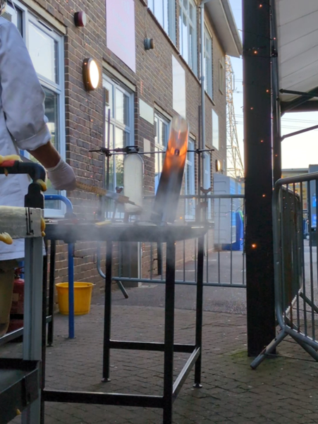 Image shows a science demonstration taking place outdoors, with a piece of equipment releasing steam and a glowing heated object held above a metal frame. A member of staff wearing protective gloves stands to the side, with the school building, safety barriers and equipment visible in the background.