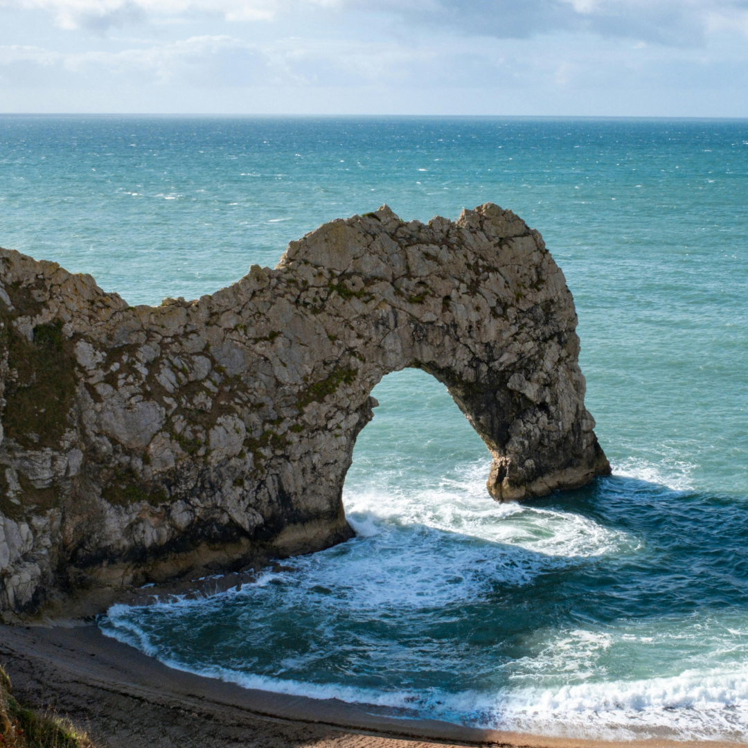 Image shows the natural limestone arch of Durdle Door extending into the sea. Waves break against the base of the rock formation, with turquoise water, a pebble beach, and a bright sky in the background.